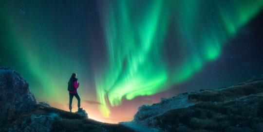 Northern lights and young woman on mountain peak at night. Aurora borealis and silhouette of alone girl on mountain trail.